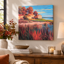Charger l'image dans la galerie, A colorful landscape canvas print of a coastal village with autumn trees and reeds, displayed on a beige wall above a wooden console table with books, a lamp, and a vase of red berries.