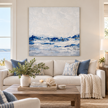 Load image into Gallery viewer, A large square abstract canvas print with blue and white ocean-like brushstrokes hangs above a cream sofa in a bright, modern living room. The room includes blue accent pillows, a wooden coffee table with books and a vase, and a window on the left showing water outside.