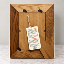 Load image into Gallery viewer, The back of a rectangular picture frame made of medium brown wood with a visible grain pattern, showing black hanging clips and a paper tag attached to the center.