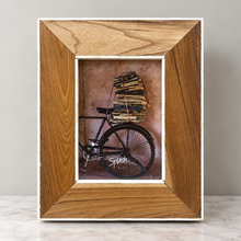 Load image into Gallery viewer, A square wooden picture frame with a natural oak finish and white inner borders, displaying a vintage-style illustration of a bicycle carrying a stack of books. The frame is photographed standing on a light marble surface against a neutral gray wall.