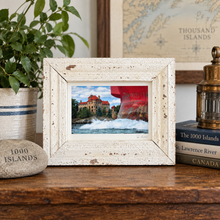 Load image into Gallery viewer, A square framed photo print with a distressed white wood frame sits on a wooden table. The photo depicts a coastal scene with blue water, white waves, a red ship, and a castle on an island.
