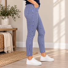 Charger l'image dans la galerie, A person wearing blue and white vertically striped cropped pants with white sneakers, standing in a bright indoor room with a wooden side table, plant, and woven rug.