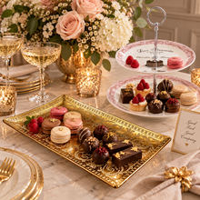 Load image into Gallery viewer, An elegant dessert spread on a marble table, featuring a large ornate gold rectangular platter with macarons and chocolates, a two-tiered white stand with macarons and pastries, champagne flutes, gold candle holders, and floral arrangements in the background.