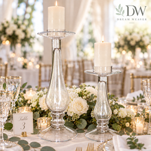 Load image into Gallery viewer, Two clear glass taper candlestick holders with lit white pillar candles, displayed on a decorated wedding table with floral centerpieces, gold candlelight holders, and tableware. The Dream Weaver logo appears in the top right corner.