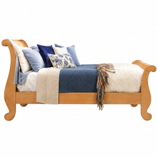 Load image into Gallery viewer, A wooden sleigh bed frame with a warm natural finish, styled with blue and cream bedding, patterned pillows, a beige knit throw, and a white faux-fur rug on a solid white background.