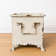 Load image into Gallery viewer, A vintage-style metal planter with a distressed off-white painted finish, featuring two U-shaped handles on the front and decorative feet, set on a light wood floor against a white wall.