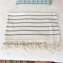 Load image into Gallery viewer, A cream-colored cotton Turkish towel with thin navy blue horizontal stripes and fringed tassels, laid flat on a white quilted bedspread.