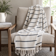 Load image into Gallery viewer, A cream throw blanket with navy blue horizontal stripes and tasseled fringe, draped over a beige armchair in a bright, modern living room setting.