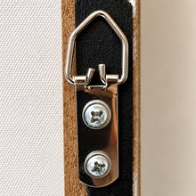 Load image into Gallery viewer, Close-up of a silver metal hanging ring and mounting bracket with two screws, attached to a black strip over a brown wooden frame, next to a white textured fabric background.
