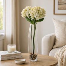 Charger l'image dans la galerie, A tall clear glass vase filled with white hydrangea stems sits on a round wooden table in a softly styled living room. Books, a candle, a small dish, and part of a sofa are visible in the background.
