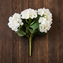 Load image into Gallery viewer, A bouquet of white faux hydrangea flowers with green leaves and stems, arranged on a dark brown wooden plank background.
