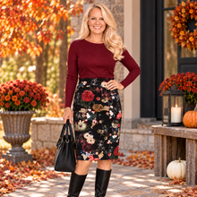Load image into Gallery viewer, A woman stands outdoors in front of a house entrance wearing a burgundy long-sleeve top and a black knee-length floral skirt with red, cream, and tan flowers. She is holding a black handbag and wearing black knee-high boots.
