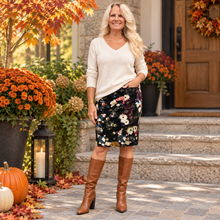 Load image into Gallery viewer, A woman stands on a stone porch wearing a cream V-neck sweater, a black floral asymmetrical skirt, and brown knee-high boots. The background features autumn decorations including orange pumpkins, potted orange flowers, and a wooden front door.
