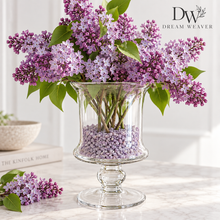 Charger l'image dans la galerie, A bouquet of purple lilac flowers arranged in a clear glass pedestal vase filled with small lavender beads, set on a light marble surface. The background is a softly blurred white interior, with a decorative book and bowl on the left and a visible 'DW Dream Weaver' logo in the top right.
