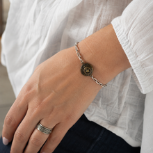 Charger l'image dans la galerie, A close-up of a person's hand and wrist wearing a silver chain bracelet with a round, textured dark bronze button charm, paired with a silver ring on the finger.
