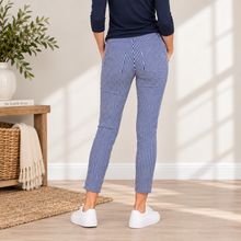 Charger l'image dans la galerie, A person shown from the back wearing blue and white vertically striped cropped pants, a navy long-sleeve top, and white sneakers in a bright indoor room with wood flooring and a woven rug.

