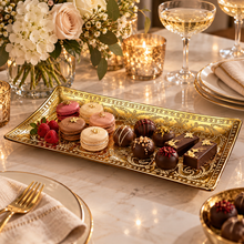 Load image into Gallery viewer, An ornate rectangular gold serving platter filled with assorted macarons, chocolate truffles, and chocolates, set on a marble table with champagne glasses, flowers, and candles in the background.

