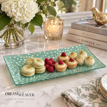 Load image into Gallery viewer, A rectangular teal glass serving platter with white polka dots is styled on a marble table, holding macarons, raspberry tarts, and white chocolate desserts. The platter is branded with the Dream Weaver logo in the lower left corner, surrounded by decorative props including a candle, books, and flowers.
