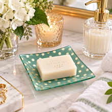 Load image into Gallery viewer, A bar of Natural Botanicals soap rests on a green glass square dish with gold polka dots, set on a marble bathroom counter with a candle, flowers, and a soap dispenser nearby.
