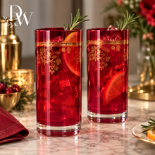 Load image into Gallery viewer, Two tall clear glasses filled with red beverage, ice, orange slices, and rosemary sprigs, set on a marble countertop with holiday decor in the background. The glasses have gold ornamental bands near the rim.
