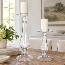 Charger l'image dans la galerie, Two clear glass candlestick holders with white pillar candles, displayed on a wooden table in a bright, modern dining or living room setting.
