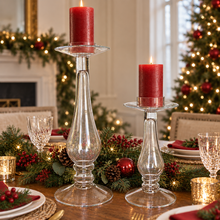 Charger l'image dans la galerie, Two clear glass candlestick holders with red pillar candles, styled on a holiday table runner with green garlands, pinecones, and a Christmas tree in the background.
