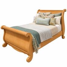 Load image into Gallery viewer, A wooden sleigh bed with a warm honey finish, styled with a sage green throw blanket, light gray and white bedding, and decorative pillows, shown on a solid white background.
