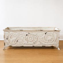 Load image into Gallery viewer, A distressed white cast iron rectangular planter box with ornate floral and scrollwork relief details and small claw feet, shown on a light wood floor against a white wall.
