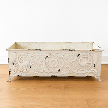 Load image into Gallery viewer, An ornate rectangular metal planter box with a distressed white finish, featuring embossed floral and scrollwork patterns and small decorative feet, set on a light wood floor against a white wall.
