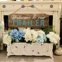 Load image into Gallery viewer, A rustic wooden sign reading 'Welcome to our TRAILER' in teal lettering, hanging above a white decorative planter filled with white and blue hydrangeas, set in front of a cream-colored cabinet.
