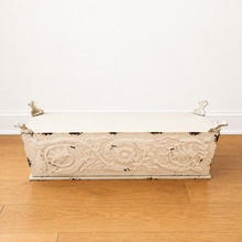 Load image into Gallery viewer, An upside down distressed cream-coloured metal planter with carved floral details and small metal handles, set on a light wood floor against a white wall.
