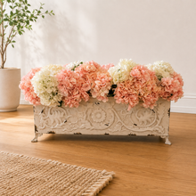 Load image into Gallery viewer, A decorative rectangular planter box with ornate embossed floral detailing and small feet, filled with faux pink and cream hydrangea flowers, placed on a wooden floor in a bright room.
