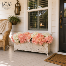 Load image into Gallery viewer, A decorative white carved rectangular planter box filled with large pink and white faux hydrangea flowers, placed on a porch beside a wicker chair, window, and black front door.
