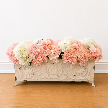 Load image into Gallery viewer, A long, ornate distressed white metal planter box with floral detailing, filled with pink and white faux hydrangea flowers, set on a light wood floor against a plain white wall.
