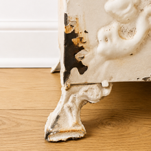 Load image into Gallery viewer, A close-up of an antique planter leg with heavily distressed, peeling white enamel and exposed rusted metal, standing on a light brown wooden floor against a white wall.
