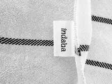 Load image into Gallery viewer, Close-up of a white woven textile with black diagonal stripe accents and a small white fabric label reading 'indaba'.
