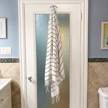 Load image into Gallery viewer, A white bathroom door with a frosted glass panel in the center, hanging a white fringed towel with thin black stripes. The bathroom has blue-gray walls, beige tiled lower walls, a white vanity on the left, and a white toilet on the right.
