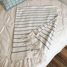 Load image into Gallery viewer, A cream-colored cotton Turkish towel with thin navy blue horizontal stripes and fringed edges, laid out on a cream-colored tufted bedspread.
