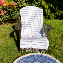 Load image into Gallery viewer, A white cotton towel with thin dark blue horizontal stripes and fringed edges, draped over a dark wooden outdoor chair on a green grass lawn with garden plants in the background.
